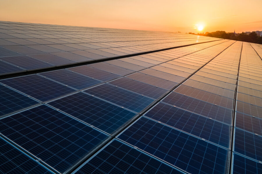 photo blue photovoltaic solar PV panels mounted on building roof for renewable energy production; sun and treetops in the background