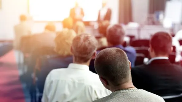 photo from conference, back of meeting room perspective shows people engaged in presentation
