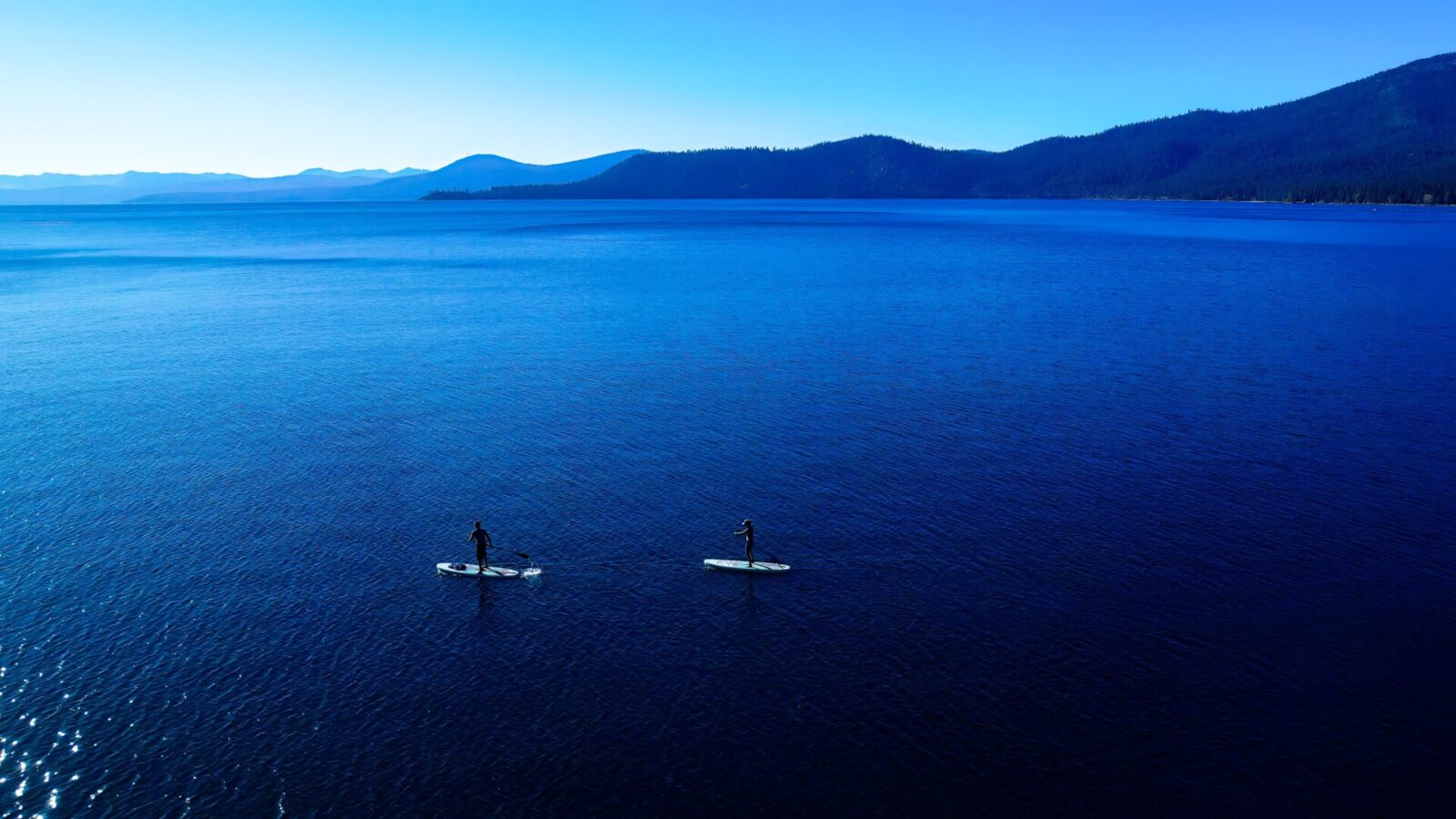 photo of blue Lake Tahoe water with stand-up paddleboarders in the foreground and mountains in the background, source Roland Schumann via unsplash