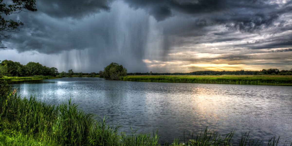 photo dark clouds and intense rainstorm in the distance; relatively calm river and vibrant green grasses in the foreground