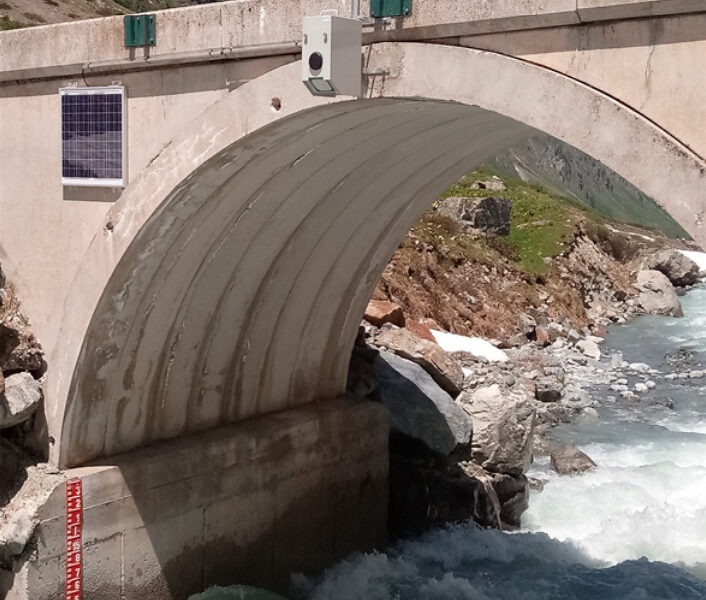 photo RQ-30 mounted on a bridge over a river; non-contact radar sensor takes discharge measurements and keeps public works workers safe. bottom of photo shows a red staff gauge to measure water level