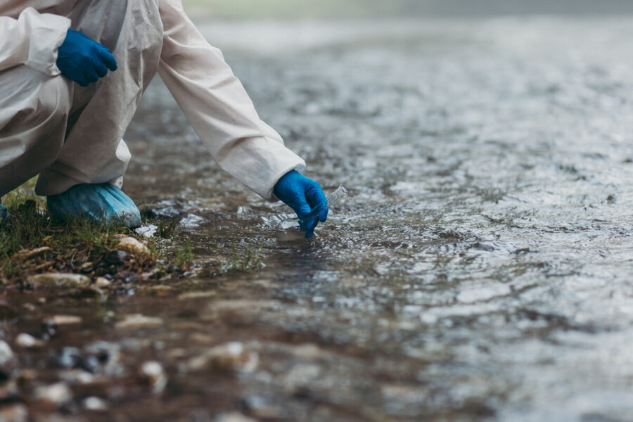 closeup photo environmental scientist collects water quality sample from surface water; gloved hand holds a glass test tube | source AdobeStock