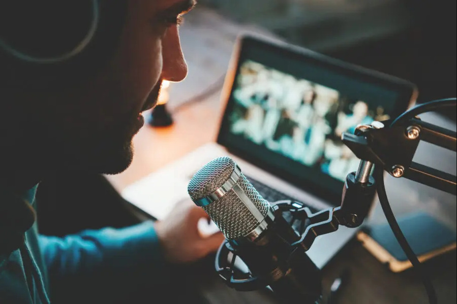 photo of a person wearing headphone, sitting in from of a microphone and a laptop to record a podcast