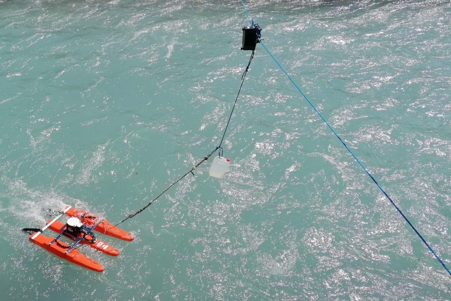 A small orange watercraft with a person aboard navigates turquoise water. A rope and pulley system suspends a plastic container above the river, possibly for scientific or environmental purposes.