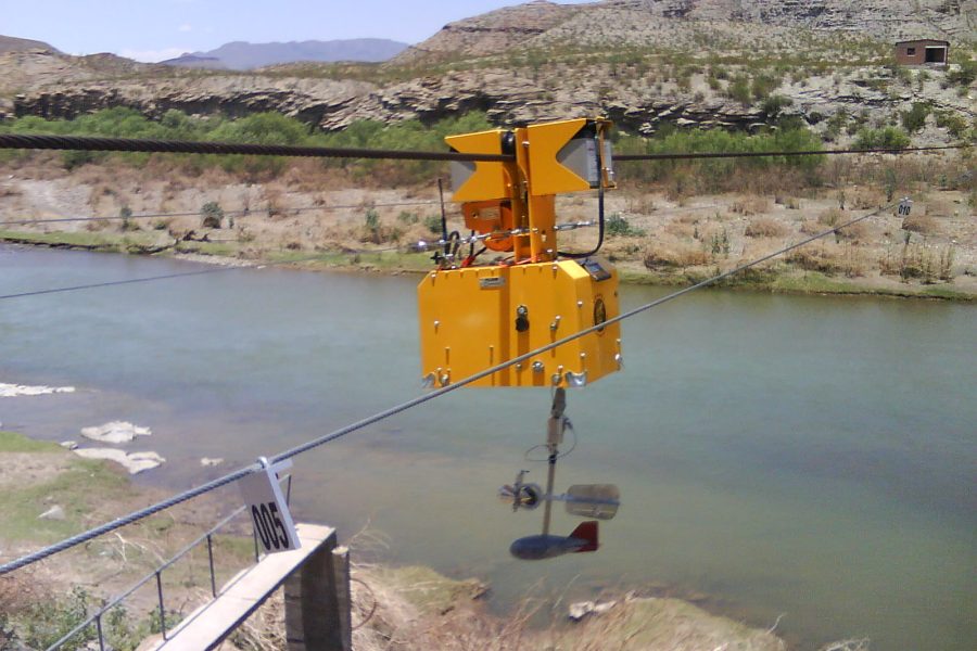 A yellow cable car-like device is suspended on a wire above a river in a rocky, arid landscape with mountains in the background. The device appears to be used for scientific or measurement purposes.