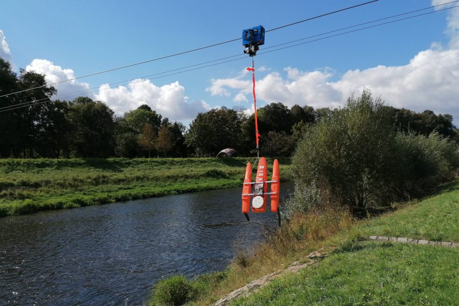 An orange watercraft is suspended above a river by a cable system, with trees and grassy banks on either side under a blue sky with scattered clouds.