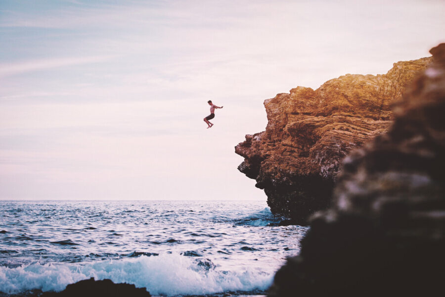 photo man jumps off cliff into water with white seafoam during the daytime