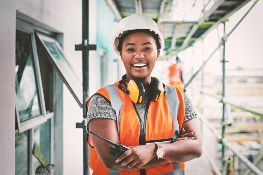 photo of a young female engineer at a renewable energy generation facility; she is wearing a hard hat, a safety vest and earphones around her neck; her arms are crossed as she holds a two-way radio and she smiles