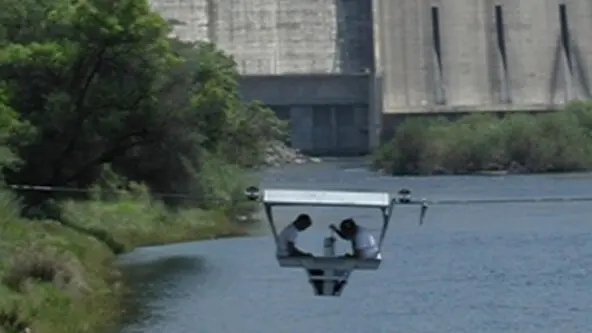photo of 2 hydrographers in gondola taking discharge measurements with hydropower dam in background